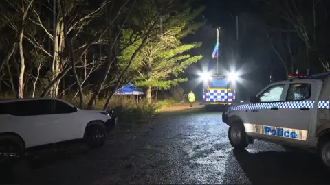 A night scene on a country road with several police cars and a small blue tent roof behind some trees.