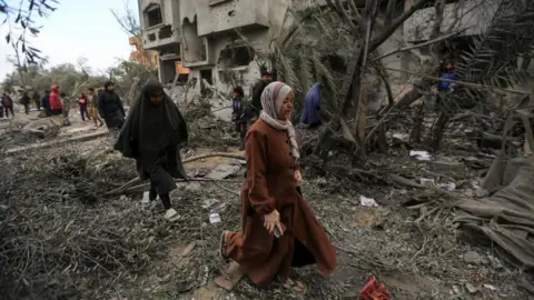 Reuters A woman walks through an area covered in debris following an air strike. A girl walks behind her and several people near a damaged house can be seen in the background. 