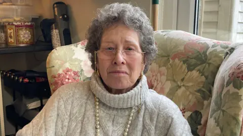A lady looking at the camera wearing glasses and a white wool jumper. She has a neutral expression. 