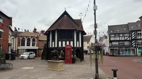 A wide shot of a town square with old dark-timbered buildings and large square cobbles