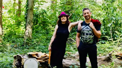 Vilk Collective Roxana Vilk with a bandmate. They are standing in a green woodland. Roxana has her arm on her bandmate's shoulder.