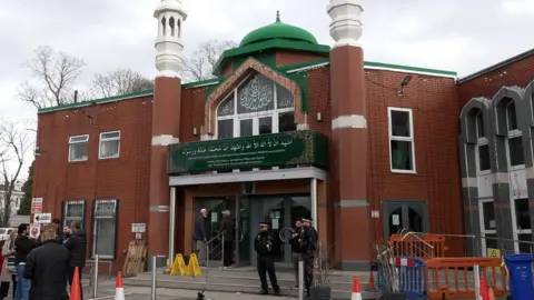 A wide shot of the Manchester Central Mosque. It is a red brick building with a green dome on top. There are multiple members of the police standing outside.