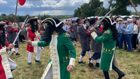 A group of men dressed as 17th century soldiers re-enact a the Battle of the Boyne. Some dressed in wither red and white or green and white uniforms are armed with swords, while some dressed in grey are armed with muskets.