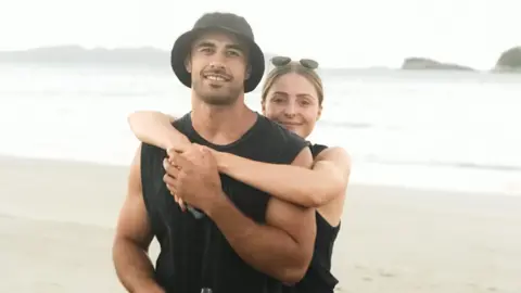 An image of a man and a woman standing on a beach. A blonde woman with sunglasses on her head has her arms around a man wearing a black vest and black bucket hat. They are both smiling at the camera.