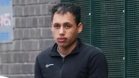 Eddie Mitchell Man in a black zipped jacket stands outside by a brick wall and metal fencing, looking toward the camera.