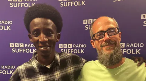 George King/BBC A young man wearing a black and white shirt with his arm around a man wearing glasses and a green top. They are both smiling and looking into the camera while standing in front of a purple BBC Suffolk backdrop.