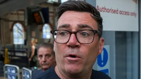 Andy Burnham pictured in the centre of the frame, wearing dark-framed glasses and a dark navy or black top. The photograph is tightly cropped around the head and shoulders. His face is sharply in focus, while most of the background is blurred. Behind him is a railway station concourse. A second man is visible in the background, but out of focus.