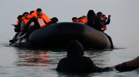 An inflatable taxi-boat filled with people approaches a person standing with their back to the camera, with water up their chest