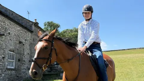 A young woman is riding a brown horse in the countryside. She is wearing a navy riding helmet, white shirt with blue spots and blue jeans. There is an old stone building in the background.