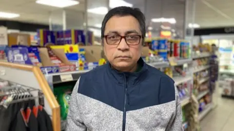 A man wearing a blue and grey zipped jumper with dark hair and glasses is standing in a convenience story with items on the shelves behind him.