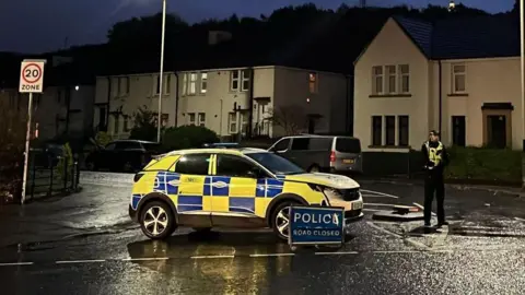 A lone police officer stands guard in the darkness on Byron Street, next to a marked police SUV, close to the scene of the fire. 