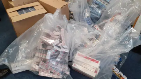 This photo shows a large amount of cigarette packs in clear plastic bags on a blue carpeted floor. Open cardboard boxes filled with more cigarette packs are in the background. The carpet they are sat on is a navy blue colour.