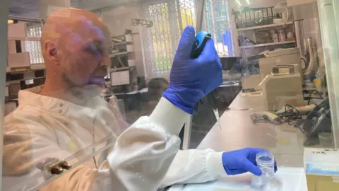 A man in a white lab coat works in a laboratory. He is moving liquid from a large pipette into a container. 