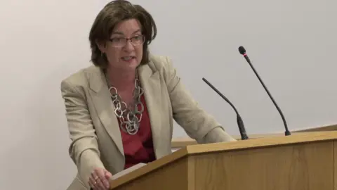 Eluned Morgan standing at a lectern answering questions from Darren Miller.