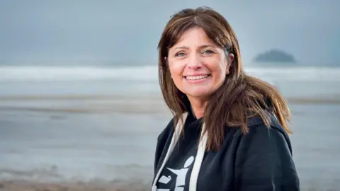 Keep Britain Tidy A brunette woman in a hoodie smiles at the camera from a beach.