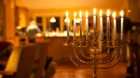 Getty Images A Chanukia, a nine-stemmed candlestick with all the flames lit in a house. In the background you can see the back of a person who is in the kitchen