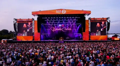 A festival stage watched by a crowd. Belinda Carlisle is performing