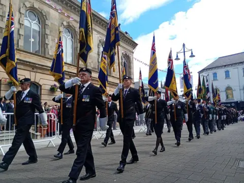 Men in military clothes, marching on a cobbled street, holding blue and yellow flags above them