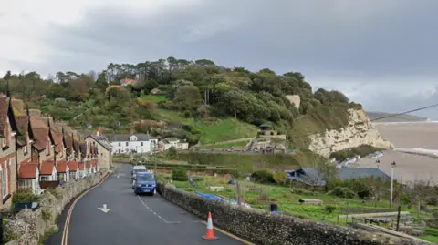 A road runs past a beach in a coastal town. A row of houses on a hill run alongside a one-way road. A small stone wall separates the road from what appears to be an allotment. Cars are parked in parking spaces on the road. Green fields and trees are on the hillside in the distance. A beach is below the cliffs.