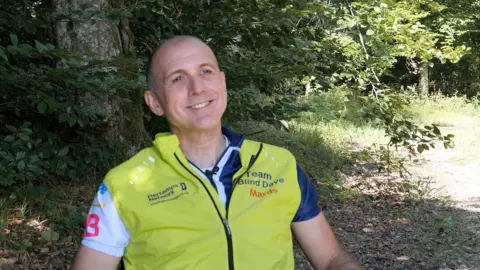 A smiling man sits in a chair with lime green gilet on with Team Blind Dave emblazoned on it 