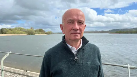 Man in green jumper stood in front of the water at Chew Valley Lake. He is bald and looking at the camera.