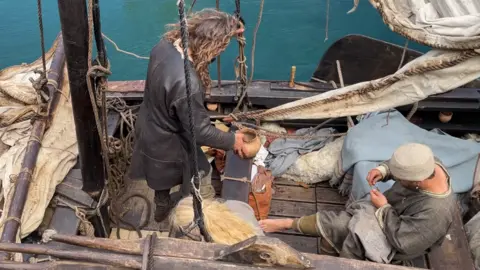 A view looking overhea onto a traditional boat. There is a man with long wavy hair standing on the left with a traditional leather jacket and holding a ceramic pot. A man on the right with woollen clothes and a woollen ht, sits barefoot sewing some fabric. The fabric of the boat's sails are around them.