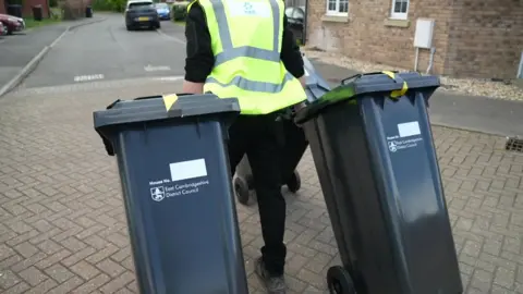 A person wearing a high visibility jacket pulls along two black wheelie bins