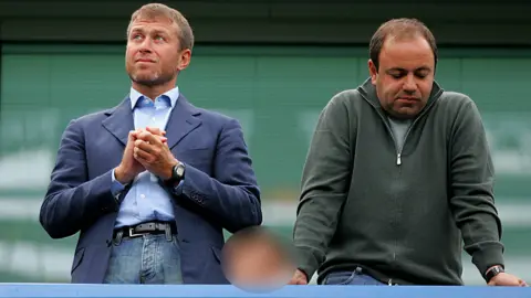 Getty Images Roman Abramovich and Eugene Shvidler standing next to each other. Mr Abramovich is wearing a blue blazer over jeans and has his hands clasped together as he looks into the distance. Mr Shvidler is wearing a quarter-zip jumper in green and is leaning on the railing, looking downwards with a resigned expression