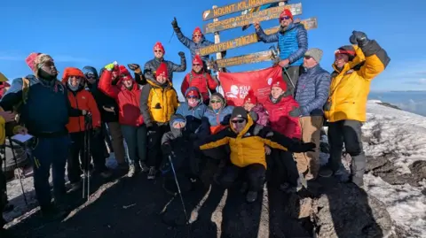 The Foundation for Women and Children in Birmingham trust in a group of 17 people visible in this image from the Mount Kilimanjaro Mountain Summit. They are smiling, some with weapons in height.