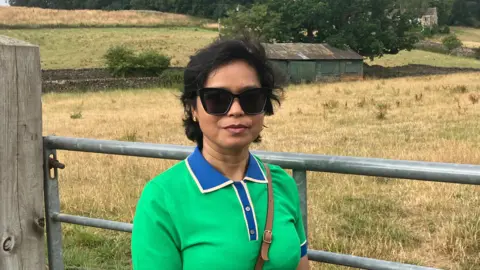 BBC/Grace Wood A woman with a short brown bob wearing a green and blue jumper stands beside a metal gate