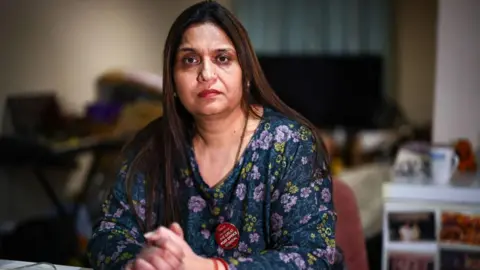 Getty Images Seema Misra looks at the camera in what looks like a home environment but which is blurred in the background. She is wearing a purple and blue flowery top and a badge which references the post office scandal.