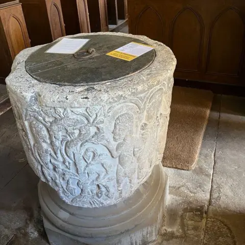 A 12th Century stone font carved with biblical figures, with a church in the background.