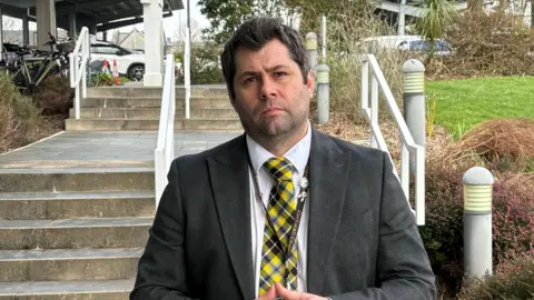 Leigh Frost wears a Cornish tartan tie, white shirt, and dak grey jacket. He has dark black hair. He stands outside the entrance to Lys Kernow with some stone steps behind him leading to the car park. 