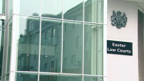 The exterior of the Exeter magistrates court. It has large glass windows that is reflecting another nearby building. To the right of the glass is a big sign that reads Exeter Law Courts in white writing on a black backdrop with a large emblem above it. 