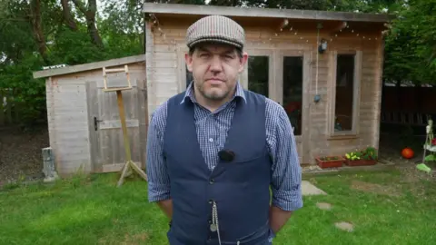 BBC Peter Chapman, wearing a waistcoat and flat cap. A wooden summer house can be seen behind him at the end of a patch of grass.