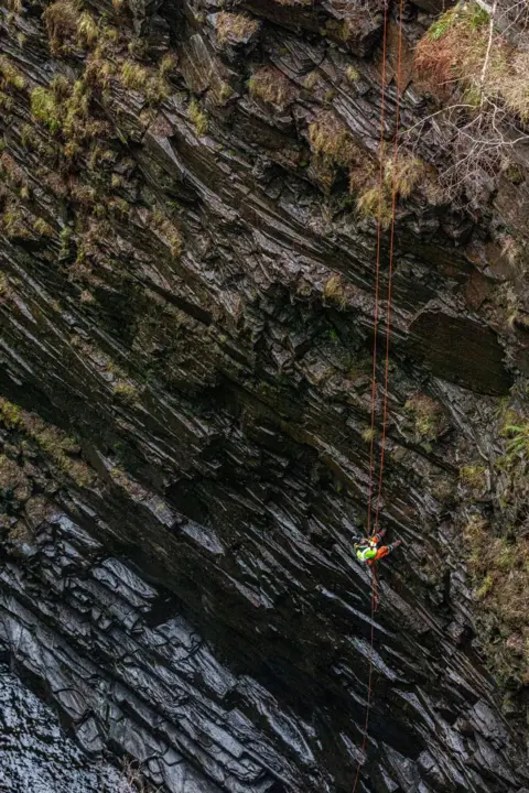 NTS A contractor wearing protective clothing and a hard hat abseils down into the gorge. The crag's rocks are dark and wet.