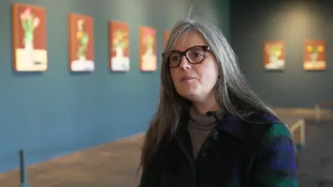 A grey-haired woman, wearing glasses and a blue and green top with a brown jumper beneath. She sits in a gallery with blue walls and rows of small paintings on display.