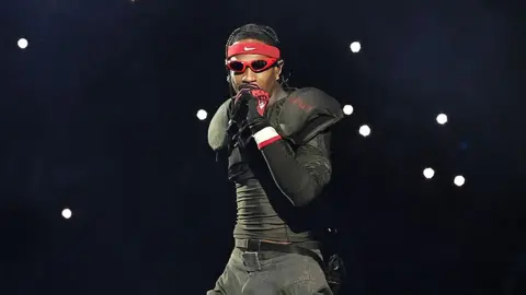 Getty Images Travis Scott, a man performing on stage, wearing all black, with red framed sunglasses and a Nike bandana on his forehead, with spotlights in the background.