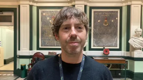 A man with brown, curly hair bushed over his forehead wearing a black jumper and lanyard with Calderdale Council insignia