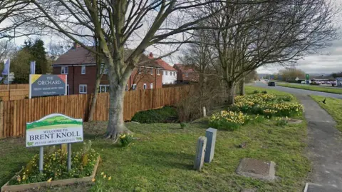 Google Images A shot showing a welcome sign for the village of Brent Knoll on a grassy verge with trees, housing behind and an A-road to the right.