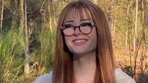 Cheshire Constabulary Brianna Ghey, with long ginger hair and wearing wide rimmed black glasses, smiles at the camera against a woodland backdrop