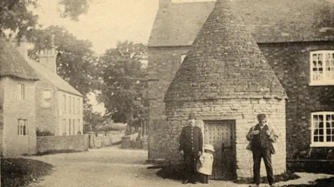 Bedfordshire Archives A black and white image of Harrold, showing the Lockup, in front of two houses, that are all made out of brick. A policeman, is standing by a door, wearing a helmet, jacket and trousers with a child next to him, wearing a white dress and hat, another man is standing to the right, wearing trousers, a jacket, waistcoat, hat and has his arms on his lapel. 