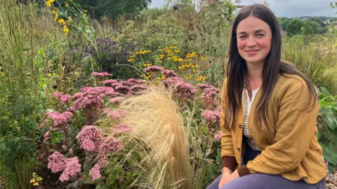 A woman with long dark hair is sitting in a garden next to some pink and yellow flours and grass.