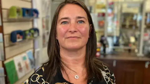 Mark Ansell/BBC Woman with shoulder length brown hair stands in front of a post office counter and looks at the camera