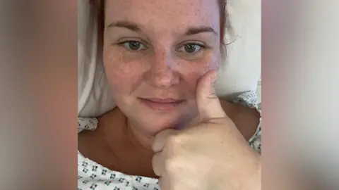 Hayley Combe/Irwin Mitchell Hayley Combe with her thumb up sat in a hospital bed