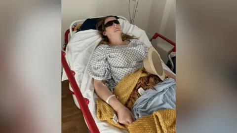 A woman lying on a hospital bed with an eye mask on and a sick bowl on top of her