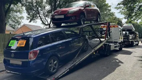 Four vehicles, on two transporters, on a road, with trees on it. There is a blue and a red vehicle on the back of the white lorry. 