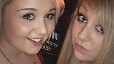 A selfie photo of two blonde girls wearing glitzy necklaces and low cut tops. They are both wearing lots of make up and smiling at the camera.