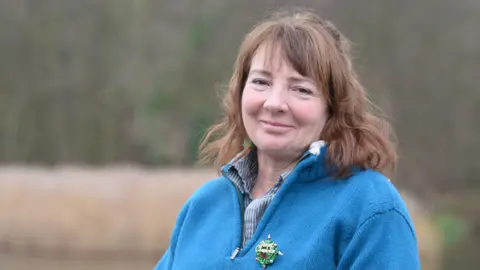 Elizabeth Dark Eilish Rothney, a reserve manager for the Norfolk Wildlife Trust. She is standing outdoors with her body turned towards the left. She is looking directly at the camera and smiling and is wearing a blue outdoor zip up fleece.