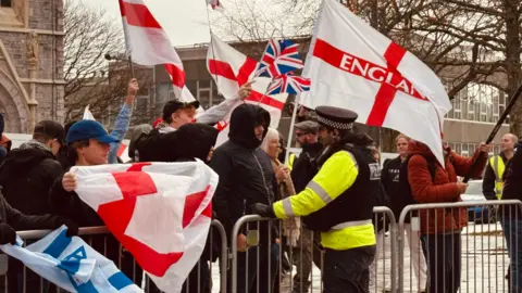 BBC A picture of people standing by a barrier. There is a man in a black coat speaking to a police officer.